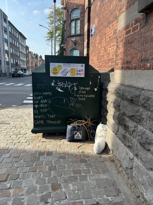 Une bulle verte de collecte de textiles avec des graffitis et des sacs de dépôts sauvages abandonnés à son pied dans une rue à Ixelles.